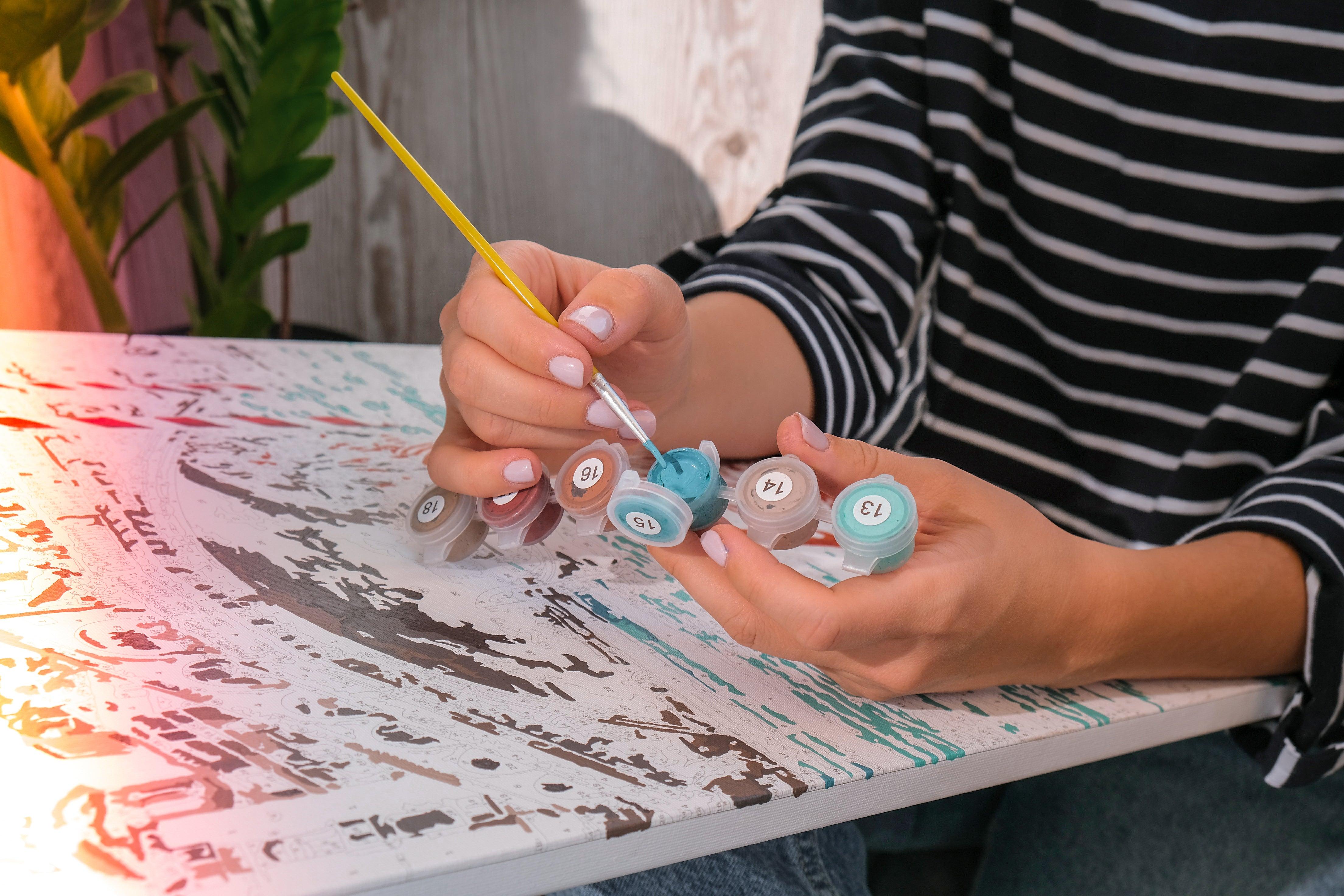 A person is sitting at a table, painting a picture using a brush and multiple paint colors.